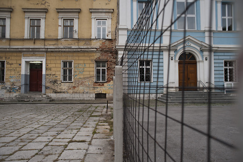 ‘Two Schools Under One Roof’ refers to the schools in Bosnia and Herzegovina, which have been ethnically divided since the Croat-Bosniak war (1992-1994).  Children from two different ethnic groups attend classes under the same roof but are physically separated from each other and taught different curicula.  At the school in the photo, the Muslim Bosniaks attend classes in the left side of the building, while the Catholic Croats attend the renovated school on the right side. 